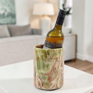 May include: A cylindrical wine cooler crafted from polished green and orange onyx stone with natural swirling patterns. A dark glass wine bottle with a black label is placed inside the stone vessel, which features a smooth, rounded rim.
