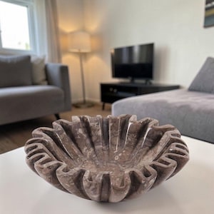 May include: A decorative, brown marble bowl with a fluted, scalloped rim. The bowl is sitting on a white table in a living room setting. The bowl's interior has a speckled pattern.
