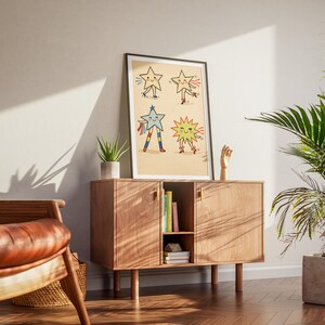 May include: A framed art print featuring four cartoon stars with arms and legs. The stars are in various poses and colors, set against a beige background. The print is displayed on a wooden cabinet with a small plant and a wooden hand sculpture.