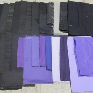 May include: An assortment of fabric swatches in various shades of black, gray, and purple. The fabrics are arranged in a layered pattern, showcasing a range of textures and hues. The collection includes solid colors and possibly some with subtle patterns.