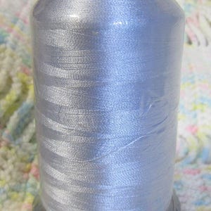 May include: A spool of light silver-grey embroidery thread wrapped in clear plastic film. The black top label features the white text ROBISON-ANTON. The thread is wound onto a black plastic base, showcasing a smooth, lustrous, and reflective finish.