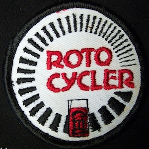 May include: A white, round embroidered patch with black and white stripes around the edge. The center of the patch features the words "ROTO CYCLER" in red embroidery. A red and white illustration of a glass of liquid is below the text.