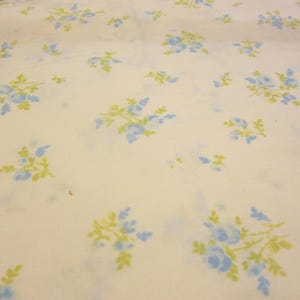 May include: Cream-colored fabric with a repeating pattern of blue and yellow floral bouquets. The flowers are clustered together with green leaves, creating a vintage aesthetic. The fabric appears to be a sheet or similar textile.