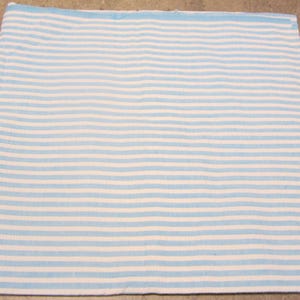 May include: A rectangular piece of fabric with horizontal stripes in light blue and white. The fabric appears to be a woven material, possibly cotton or linen, and is laid flat on a neutral surface.