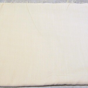 May include: A rectangular piece of off-white fabric with a smooth, woven texture. The material features visible, slightly frayed edges along the sides, indicating a raw cut. The color is a consistent, light cream shade throughout the entire surface.