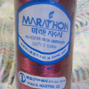 May include: A spool of maroon embroidery thread, labeled "MARATHON" with Korean text. The label indicates it's 100% polyester, 120/2, and 5,000m. The spool is wrapped in clear plastic with a blue top and base. "DONG-IL INDUSTRIAL CO." is also printed.