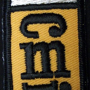 May include: A rectangular embroidered patch featuring a gold background with the black text "cm" and a black dot below it. The patch has a white horizontal bar at the top and is bordered by a thick black stitched edge on all four sides.