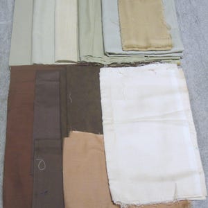 May include: A collection of fabric swatches in various neutral colors, including shades of brown, beige, and green. The fabric pieces are rectangular and appear to be made of linen or a similar material. Some swatches are folded or stacked.