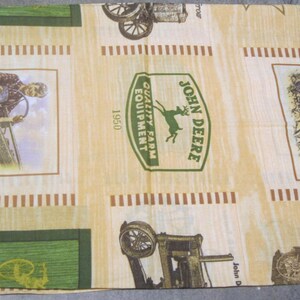 May include: A rectangular piece of fabric featuring a repeating John Deere print on a light tan background. The pattern includes green logos with a deer, the text John Deere Quality Farm Equipment 1950, illustrations of vintage tractors, and a person operating farm machinery.