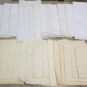 May include: A collection of rectangular fabric pieces arranged in two distinct rows. The top row consists of multiple folded sheets of bright white fabric, while the bottom row features several folded sheets of off-white or cream-colored fabric.