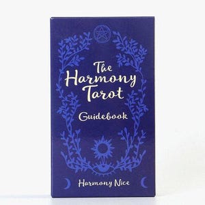 May include: A navy blue tarot guidebook titled "The Harmony Tarot Guidebook." The cover features white script and a decorative blue floral border. Additional design elements include a sun, an eye, and the name "Harmony Nice."