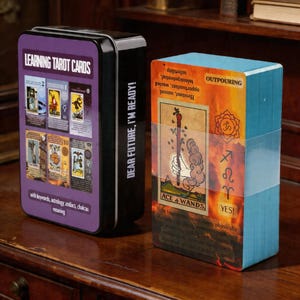 May include: A tarot card deck and storage tin. The black tin has a purple label with "LEARNING TAROT CARDS" and "DEAR FUTURE, I'M READY!". The deck is wrapped in clear plastic and features the "ACE + WANDS" card.