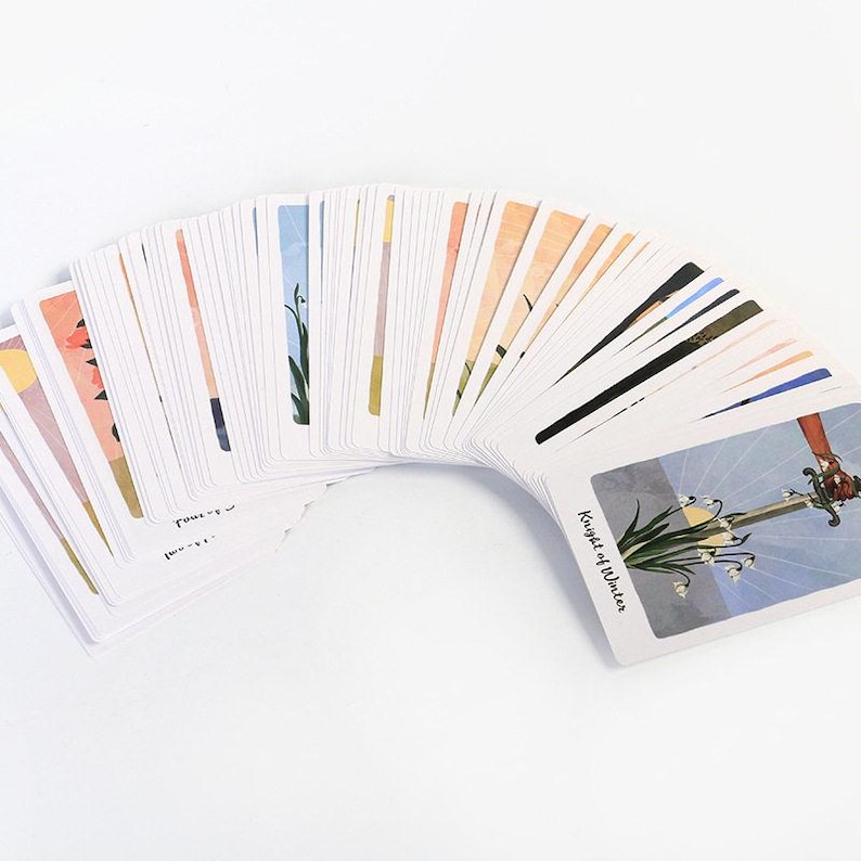 May include: A deck of tarot cards fanned out, showcasing colorful illustrations. The cards feature various images, including a sword and flowers, with the words "Knight of Winter" visible on one card. The cards are arranged on a white surface.