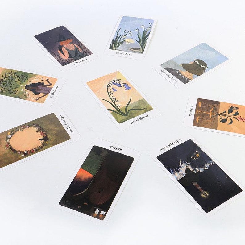 May include: A collection of tarot cards featuring watercolor illustrations. The cards display various scenes, including a witch, flowers, and abstract imagery. The cards are arranged on a white surface, showcasing their artistic designs.