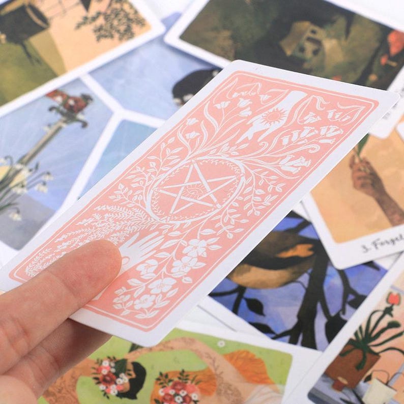 May include: A tarot card with a pink floral design and a white pentagram. The card is held in a hand, with other cards visible in the background. The cards have illustrations and text.