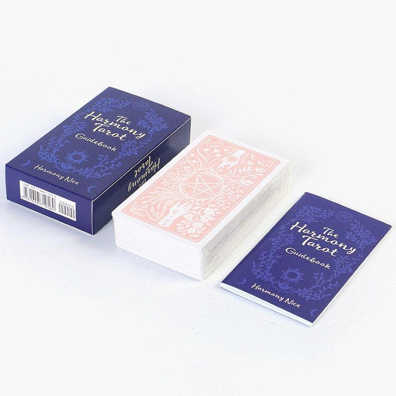 May include: A tarot card set with a navy blue box and guidebook, both featuring the text "The Harmony Tarot" and floral designs. The deck of cards has pink backs with a pentagram and hand illustration.