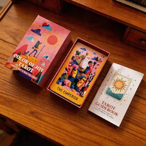 May include: A tarot card deck with a pink box labelled "COLOR OF JOY TAROT" and a card displaying "THE EMPEROR". A guidebook with a sun illustration is also present. The deck is displayed on a wooden surface.
