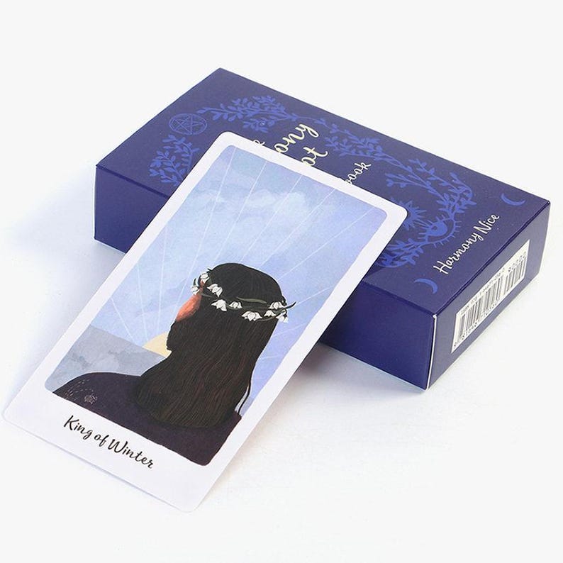 May include: A tarot card with the words "King of Winter" and an illustration of a person wearing a floral crown, resting on a blue box with the words "Harmony Nice". The card is light blue and white.