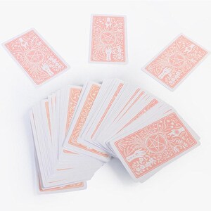 May include: A deck of tarot cards with a white and peach color scheme. The card backs feature a floral design with a pentagram and hands. Several cards are fanned out, with others scattered around.