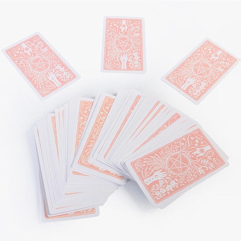 May include: A deck of tarot cards with a white and peach color scheme. The card backs feature a floral design with a pentagram and hands. Several cards are fanned out, with others scattered around.