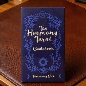 May include: A navy blue tarot guidebook titled "The Harmony Tarot Guidebook." The cover features a white script font and blue floral and celestial illustrations, including a pentagram, sun, moon, and eye. The text "Harmony Nice" is at the bottom.