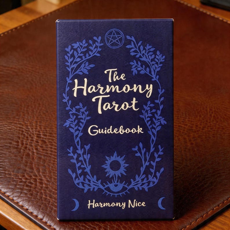 May include: A navy blue tarot guidebook titled "The Harmony Tarot Guidebook." The cover features a white script font and blue floral and celestial illustrations, including a pentagram, sun, moon, and eye. The text "Harmony Nice" is at the bottom.