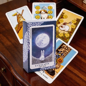 May include: A tarot deck with a blue box featuring a white rabbit under a full moon. Several tarot cards are fanned out, each with unique illustrations of rabbits and symbols. The deck is labelled "Lunalapin Tarot Deck".