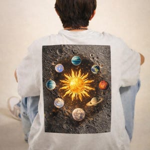 May include: White t-shirt featuring a vibrant graphic of the sun surrounded by planets on a dark, textured background. The design includes a bright yellow sun and various colorful planets, creating a celestial theme. The shirt is a casual, everyday wear item.