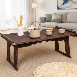 Puede incluir: Una mesa baja de madera marrón oscuro con borde festoneado, que contiene un juego de té. El juego incluye una tetera beige, dos tazas blancas pequeñas y un recipiente blanco con utensilios de madera. La mesa está en un salón.