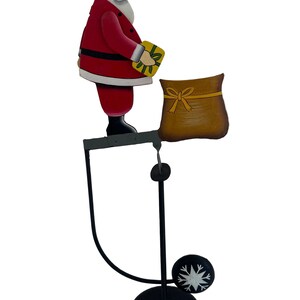 May include: A metal Santa Claus figure standing on a horizontal bar, holding a yellow gift box. A brown sack sits on the other end of the bar. The structure is supported by a thin black metal stand with a circular base and a small snowflake accent.