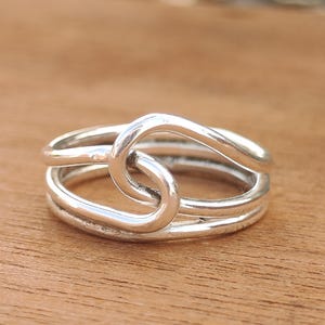 Puede incluir: Un anillo de plata con un diseño de nudo único. El anillo presenta tres bandas que se entrelazan para formar un nudo central. El metal pulido refleja la luz, creando un accesorio brillante y llamativo.