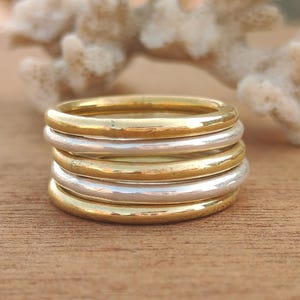 May include: A stack of five rings, alternating between gold and silver tones. The rings are smooth and simple, with a rounded profile. The background features a piece of coral and a wooden surface.