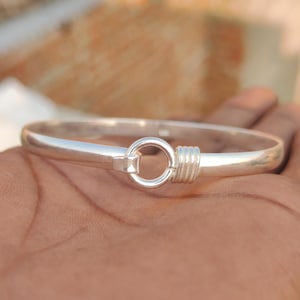 May include: A silver bangle bracelet with a circular clasp and a wrapped detail. The bracelet is smooth and shiny, reflecting light. The clasp is a simple, elegant design. The bracelet is shown on a hand.