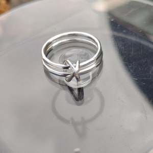 May include: A silver ring featuring three bands, with a small X-shaped design crossing one of them. The ring is highly reflective, and the image is taken in natural light, creating a shadow.