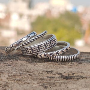 Puede incluir: Una colección de anillos de plata con varios patrones decorativos. Los anillos presentan diseños como formas geométricas repetidas, motivos de corazón y bandas texturizadas. Los anillos se muestran sobre una superficie de madera.