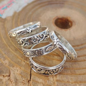 Puede incluir: Un conjunto de anillos de plata con diseños florales y de volutas. Los anillos presentan patrones detallados de flores y hojas. Se exhiben sobre una superficie de madera, mostrando su artesanía y estilo.