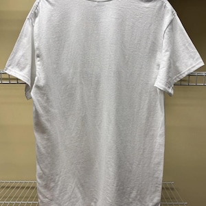 Pu&ograve; includere: Una t-shirt bianca a maniche corte con scollo rotondo. Il capo &egrave; appeso a una gruccia nera e mostra il retro della maglietta. Il tessuto appare come un materiale bianco solido e opaco con cuciture standard lungo le maniche e l'orlo inferiore.