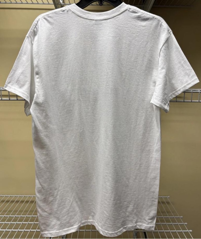 Pu&ograve; includere: Una t-shirt bianca a maniche corte con scollo rotondo. Il capo &egrave; appeso a una gruccia nera e mostra il retro della maglietta. Il tessuto appare come un materiale bianco solido e opaco con cuciture standard lungo le maniche e l'orlo inferiore.