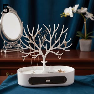 May include: A white jewellery organiser shaped like a tree with a deer figurine at the base. The organiser has branches for hanging necklaces and earrings, and a drawer for storage. The base is an oval shape.