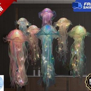 Op de afbeelding: Een cluster van zeven hanglampen in de vorm van kwallen, in verschillende kleuren zoals roze, paars en teal. Elke lamp heeft een doorschijnend, vloeiend stoffen lichaam met interne verlichting, wat een zachte, etherische gloed creëert. De lampen hangen aan het plafond.