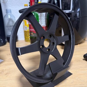 Könnte beinhalten: Ein schwarzer Sechs-Speichen-Rad-Ausstellungsständer. Der Ständer ist für ein Modellautorad konzipiert. Das Rad hat einen kreisförmigen Felgenrand und eine zentrale Nabe mit mehreren Schraubenlöchern. Der Ständer besteht aus mattem, schwarzem Material.