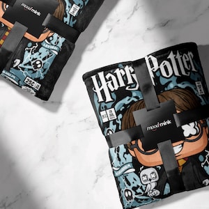 Könnte beinhalten: Zwei schwarze Decken mit Harry-Potter-Motiv, mit einer Cartoon-Illustration der Figur. Die Decken sind mit schwarzem Band und einem moodmink-Etikett umwickelt. Das Design enthält den Text "Harry Potter" und andere Grafiken.