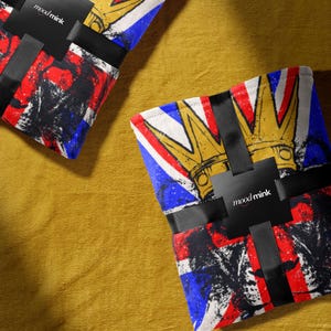 Pode incluir: Duas mantas dobradas com um design Union Jack, apresentando uma coroa dourada e elementos abstratos vermelhos, pretos e azuis. Cada manta é embrulhada com uma faixa preta e uma etiqueta que diz "mood/mink". As mantas estão sobre uma superfície dourada.