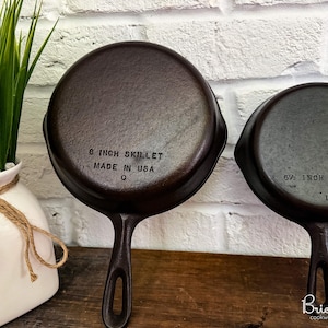 May include: Two cast iron skillets, an 8-inch (approx. 20 cm) and a 6.5-inch (approx. 16.5 cm), with long handles. The 8-inch skillet is marked "MADE IN USA". The skillets are dark brown and sit on a wooden surface. A white vase with greenery is in the background.