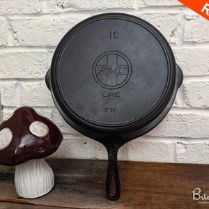 Op de afbeelding: Een zwarte Griswold gietijzeren koekenpan, gemarkeerd met "10" en "716", met het Griswold-logo. De koekenpan heeft een lange handgreep en twee zijhandgrepen. Een keramische paddenstoel siert de scène. Een rode banner leest "RARE!"