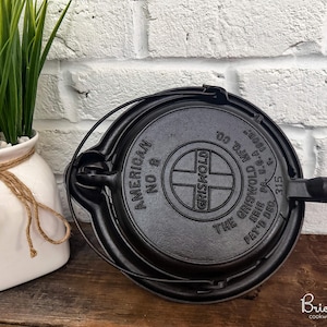 Könnte beinhalten: Schwarzes Gusseisen-Waffeleisen Griswold Nr. 8. Das Eisen hat eine runde Form mit Griff und Klappdeckel. Der Text "American No. 8" und "Griswold" ist sichtbar. Eine weiße Vase mit grünen Pflanzen befindet sich im Hintergrund.