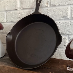 Könnte beinhalten: Eine dunkelbraune gusseiserne Bratpfanne mit langem Griff. Die Pfanne ist rund mit einer leicht abgeschrägten Seite. Das Bild enthält auch zwei dekorative Pilzfiguren und den Text "Bricksworth cookware restoration".