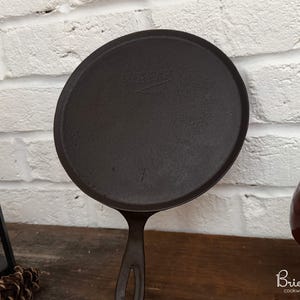 Peut inclure: Une poêle à griller en fonte de couleur marron foncé avec une longue poignée. La poêle est ronde avec une surface de cuisson plate. L'arrière-plan comprend un mur de briques blanches et une surface en bois.