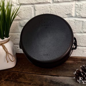 以下が含まれることがあります： 黒い鋳鉄製のWagner Ware Sidney-Oダッチオーブン、蓋付き。鍋には2つのハンドルがあり、蓋には「Wagner Ware Sidney -O-」の文字がエンボス加工されています。「1268 J」の数字が底に刻印されています。左側には緑の植物が入った白い花瓶があります。