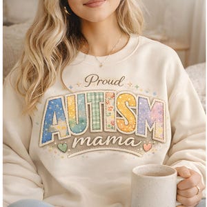 May include: Cream-colored sweatshirt with the words "Proud Autism Mama" in a colorful, watercolor-style design. The word "AUTISM" is in a rainbow gradient, and "mama" is in a simple font. The sweatshirt has a crew neck and long sleeves.